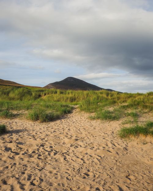 Free Tranquil scene of sandy dunes leading to lush greenery and hills under a cloudy sky in Ireland. Stock Photo