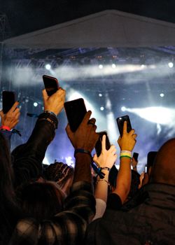 A lively concert scene with a focused crowd raising phones to film the stage. High energy and excitement.