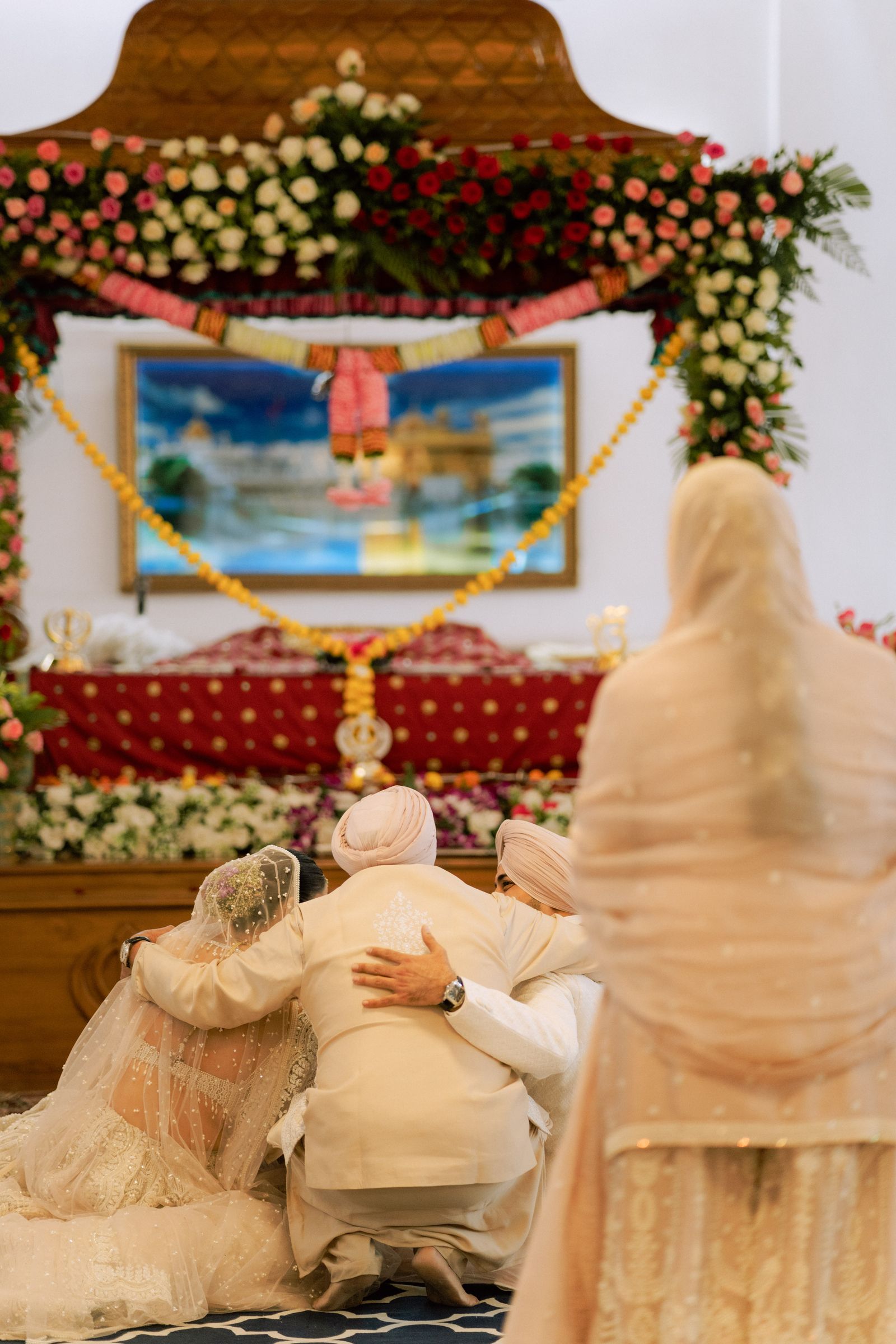 A couple kneeling before the Guru Granth Sahib under a floral palki, an elder leaning in to bless them.