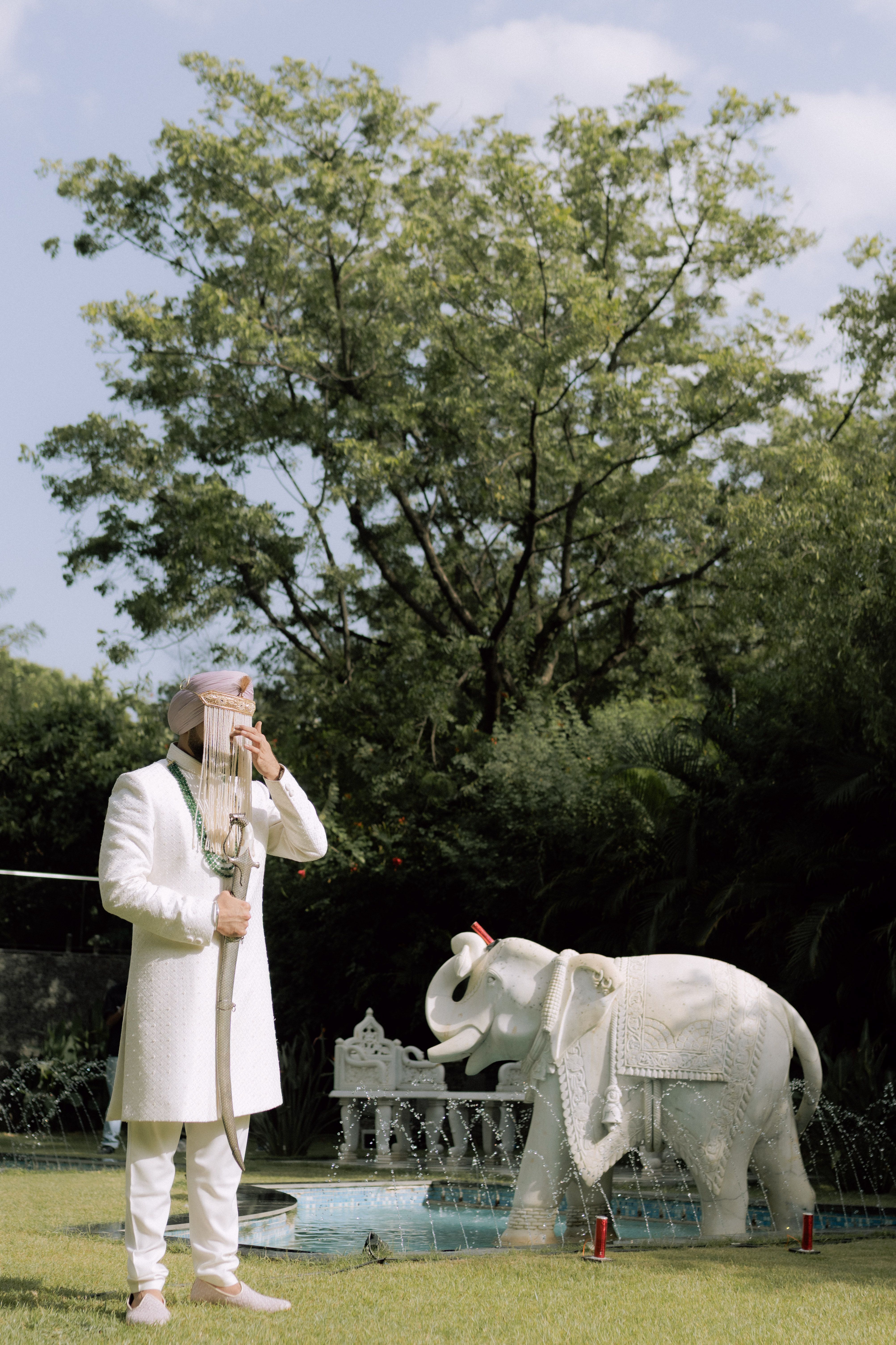 Man in traditional wedding attire beside decorative elephant statue outdoors.