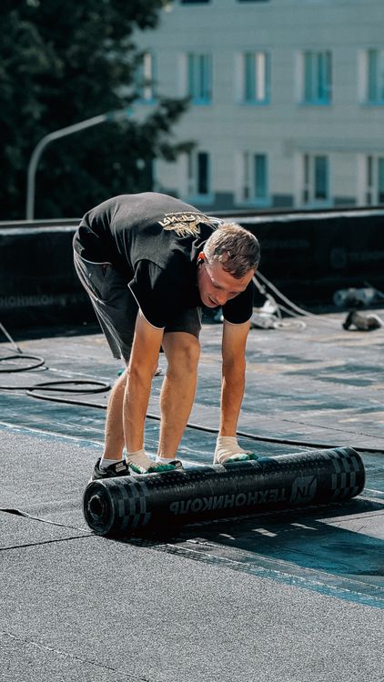 Worker rolling a waterproof membrane across a flat roof under a clear sky