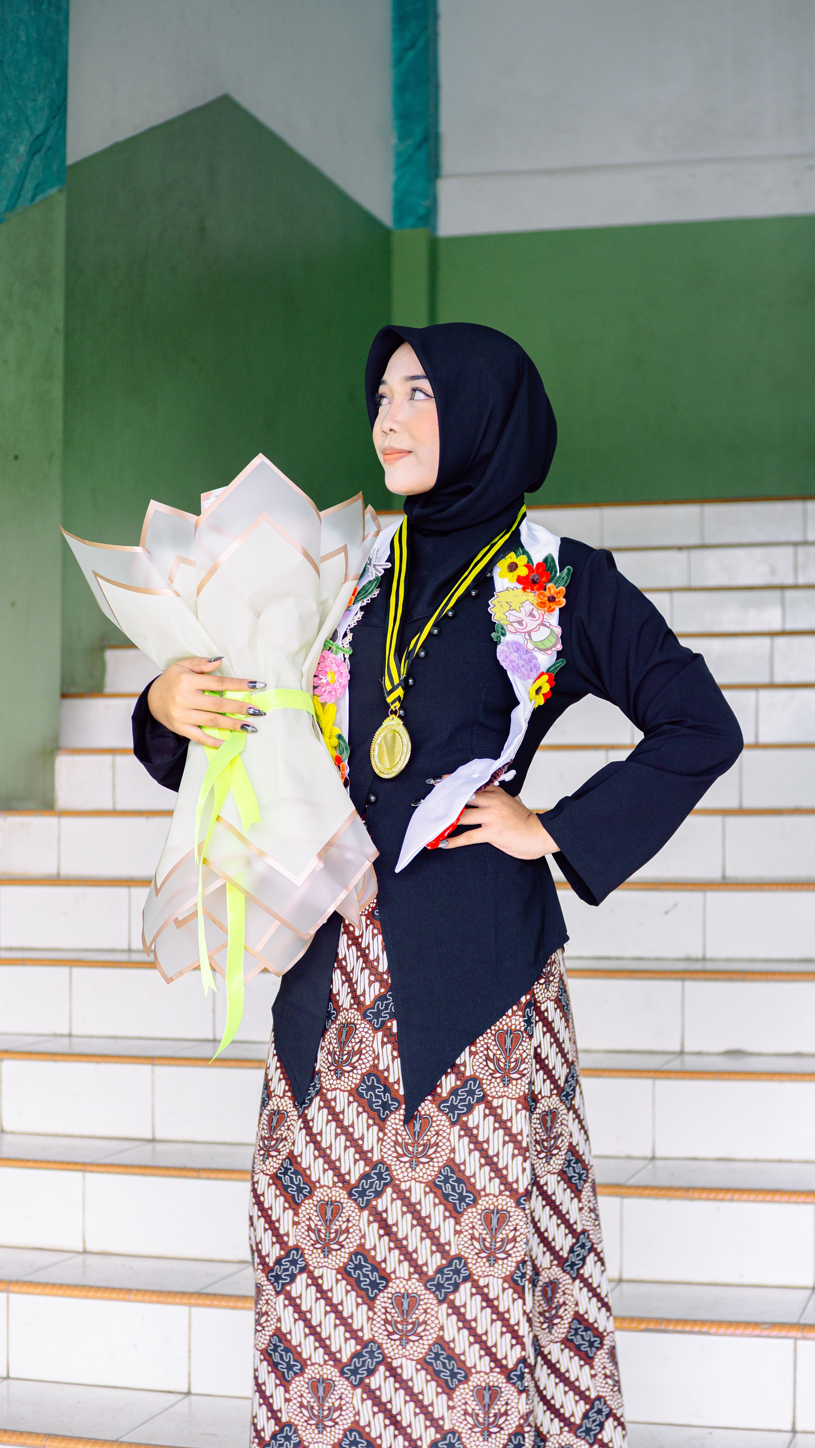 A proud graduate in traditional attire holding a bouquet on a staircase.