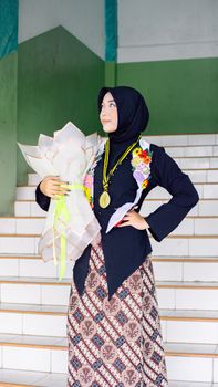 A proud graduate in traditional attire holding a bouquet on a staircase. - Fotografer Sidoarjo