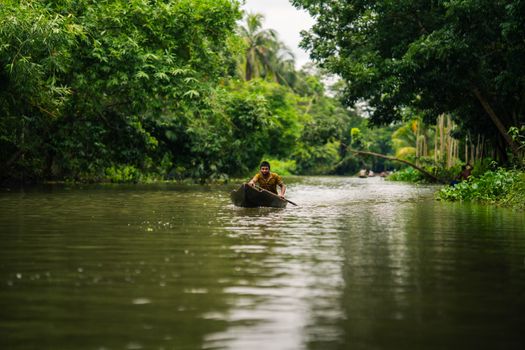 A man paddling a canoe on a calm river surrounded by lush green trees in Rajshahi, Bangladesh.