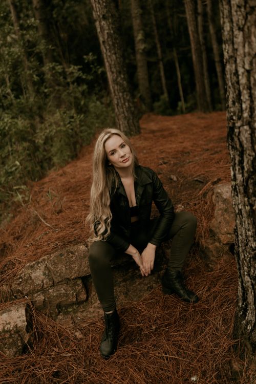 Free A woman in a black leather jacket sits in a forest, surrounded by pine needles and tall trees. Stock Photo
