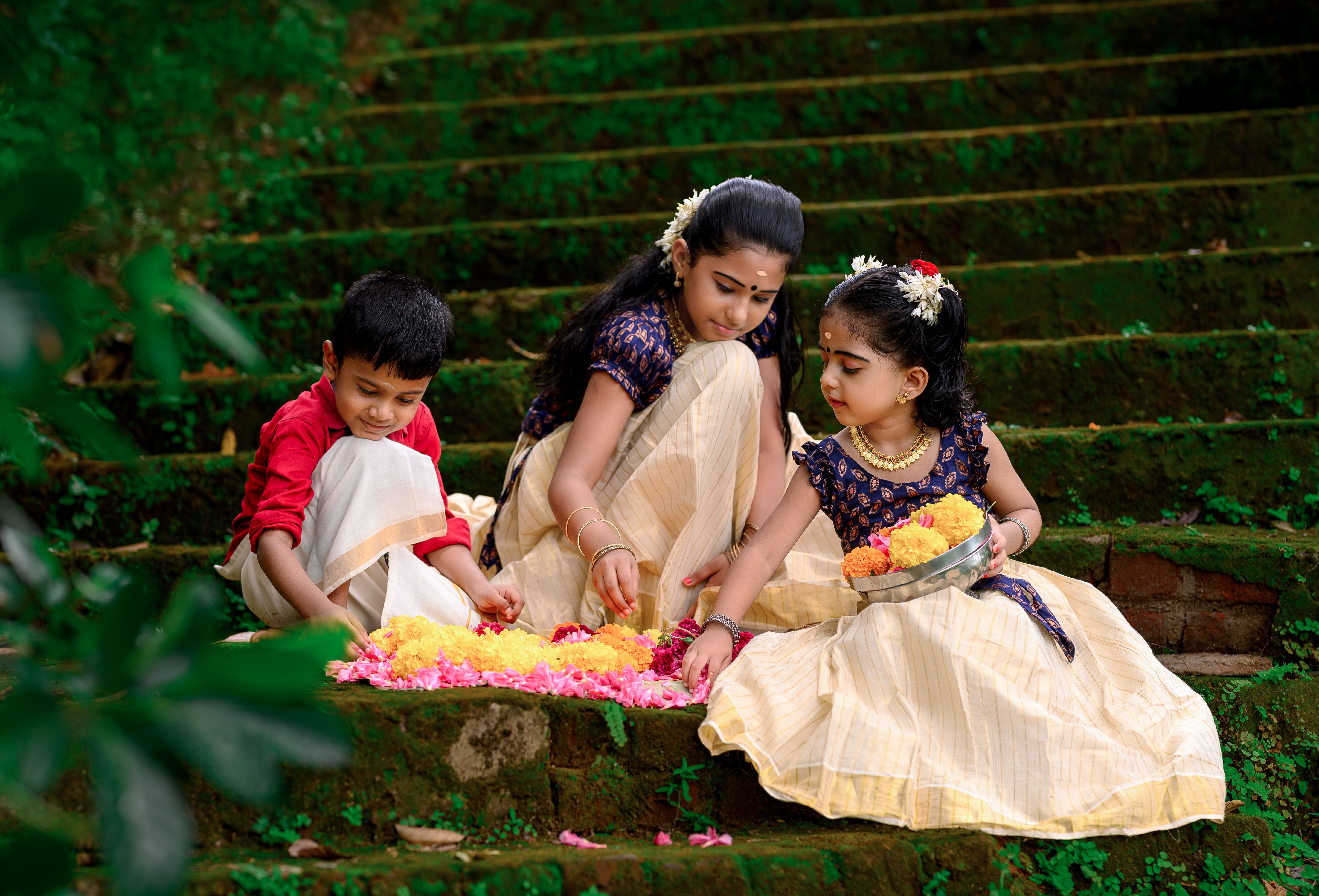 Three children in traditional attire create a flower design for Onam on stone steps in Kerala, India.