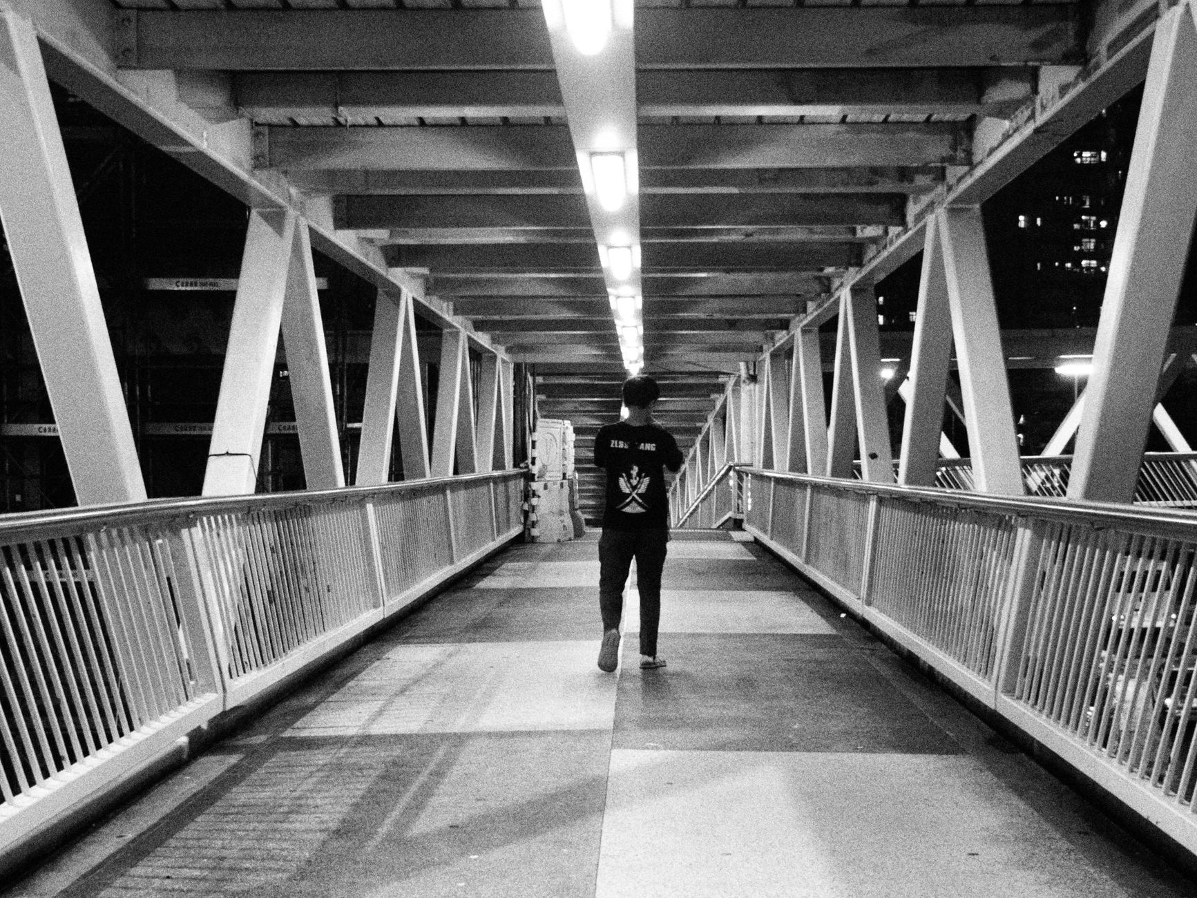 Black and white photo of a person walking alone on a city footbridge in Hong Kong at night.
