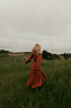 A woman in a flowing red dress twirls in a grassy field under a cloudy sky.