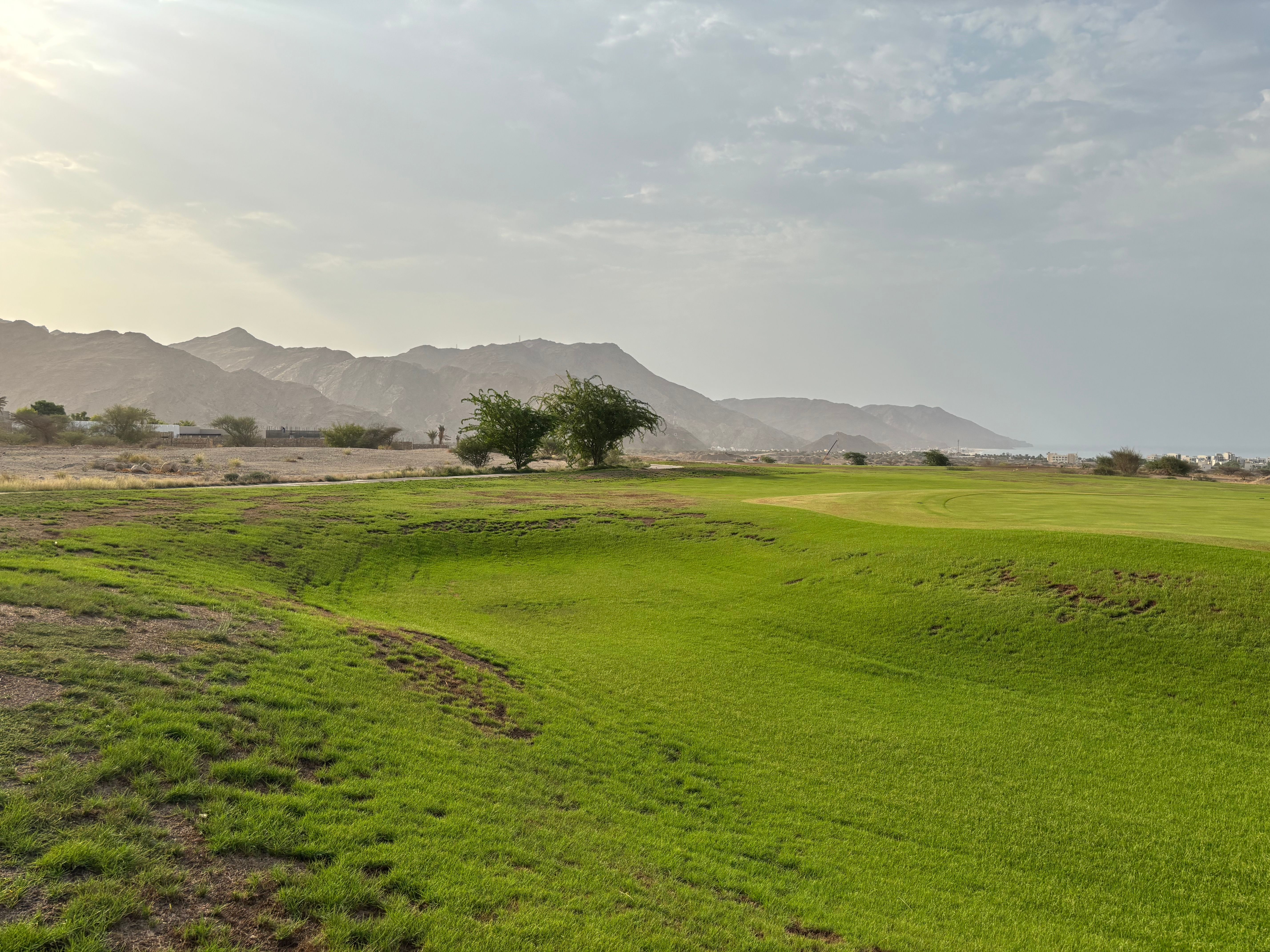Bright green golf course amidst a mountainous desert landscape under a cloudy sky.