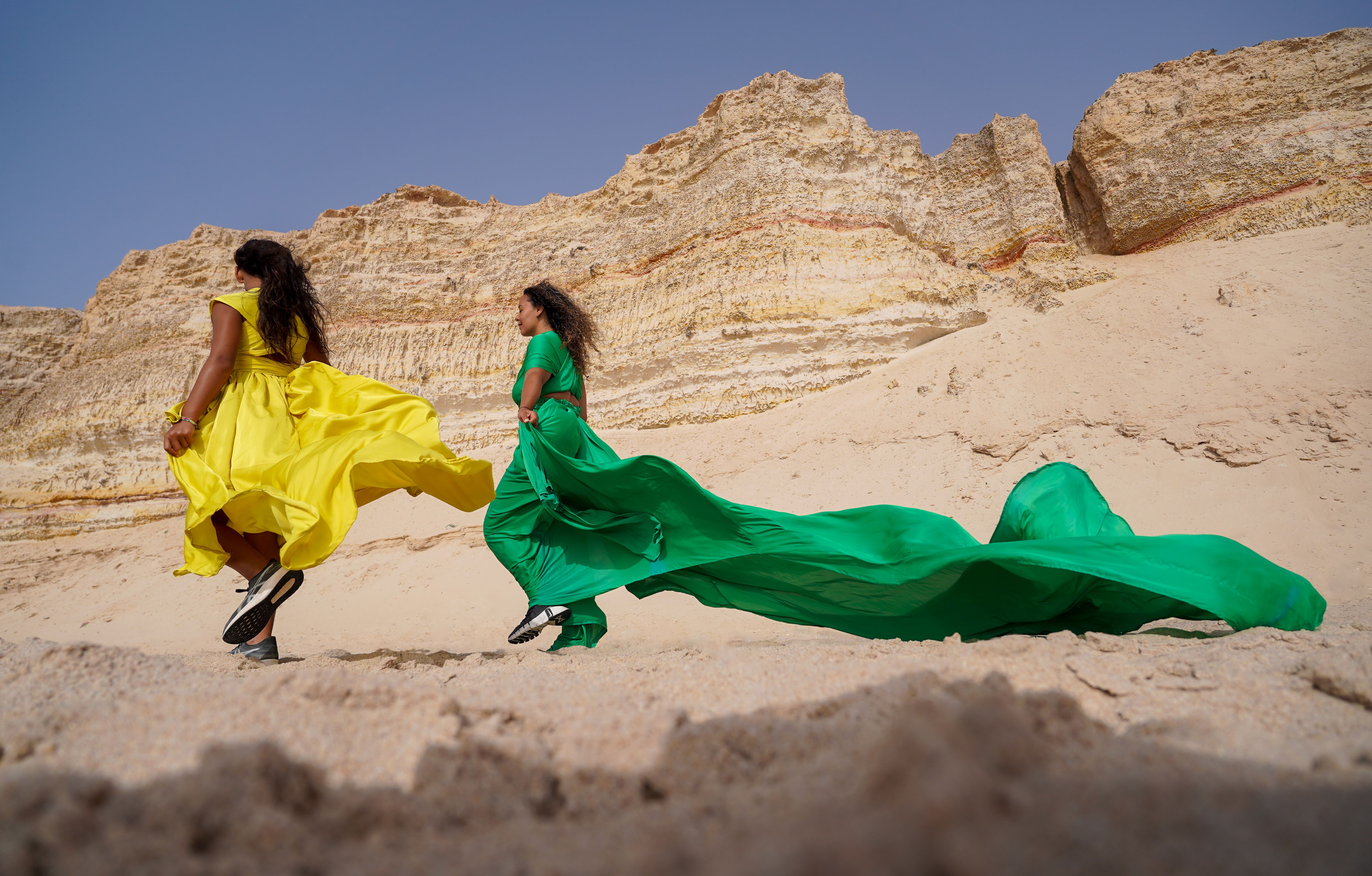 Two women in colorful dresses run across a sandy desert landscape, capturing movement and freedom.