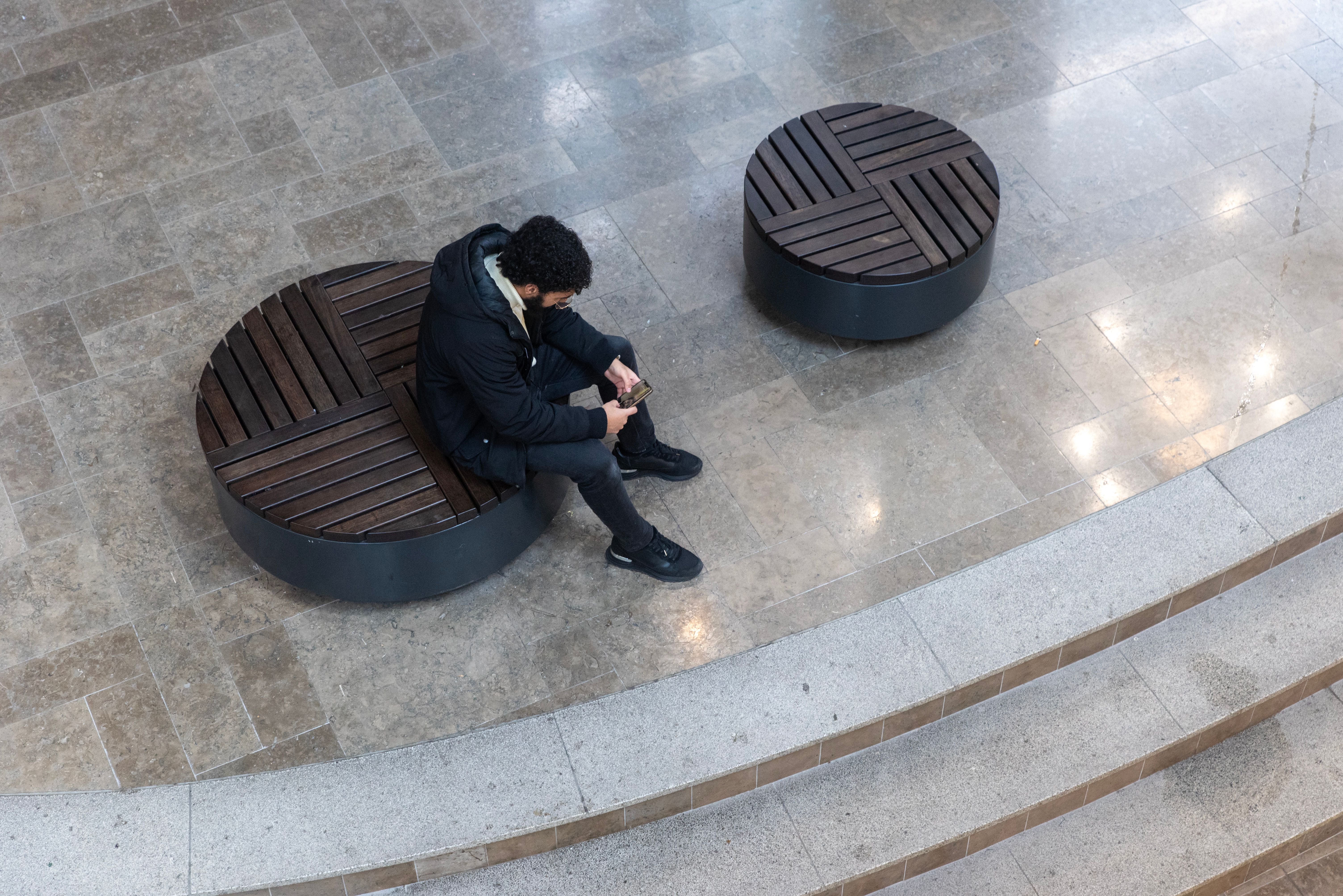 Young man sitting alone on a circular bench indoors, checking his phone.