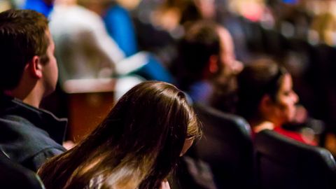 A focused shot of a person reading amidst a large audience in an indoor auditorium.