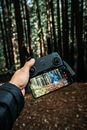 Crop unrecognizable male in padded jacket standing with reached arm with cellphone fixed to UAV remote controller holder demonstrating screen with bright picture in forest on blurred background
