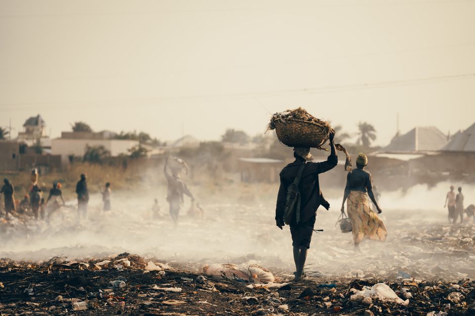 People walk through waste in a smoke-filled landscape in Burundi, highlighting pollution.