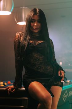 Elegant woman in black lace dress posing confidently by a pool table with soft ambient lighting.