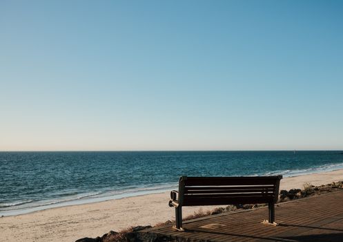Serene view of an empty bench facing a tranquil ocean under a clear blue sky.