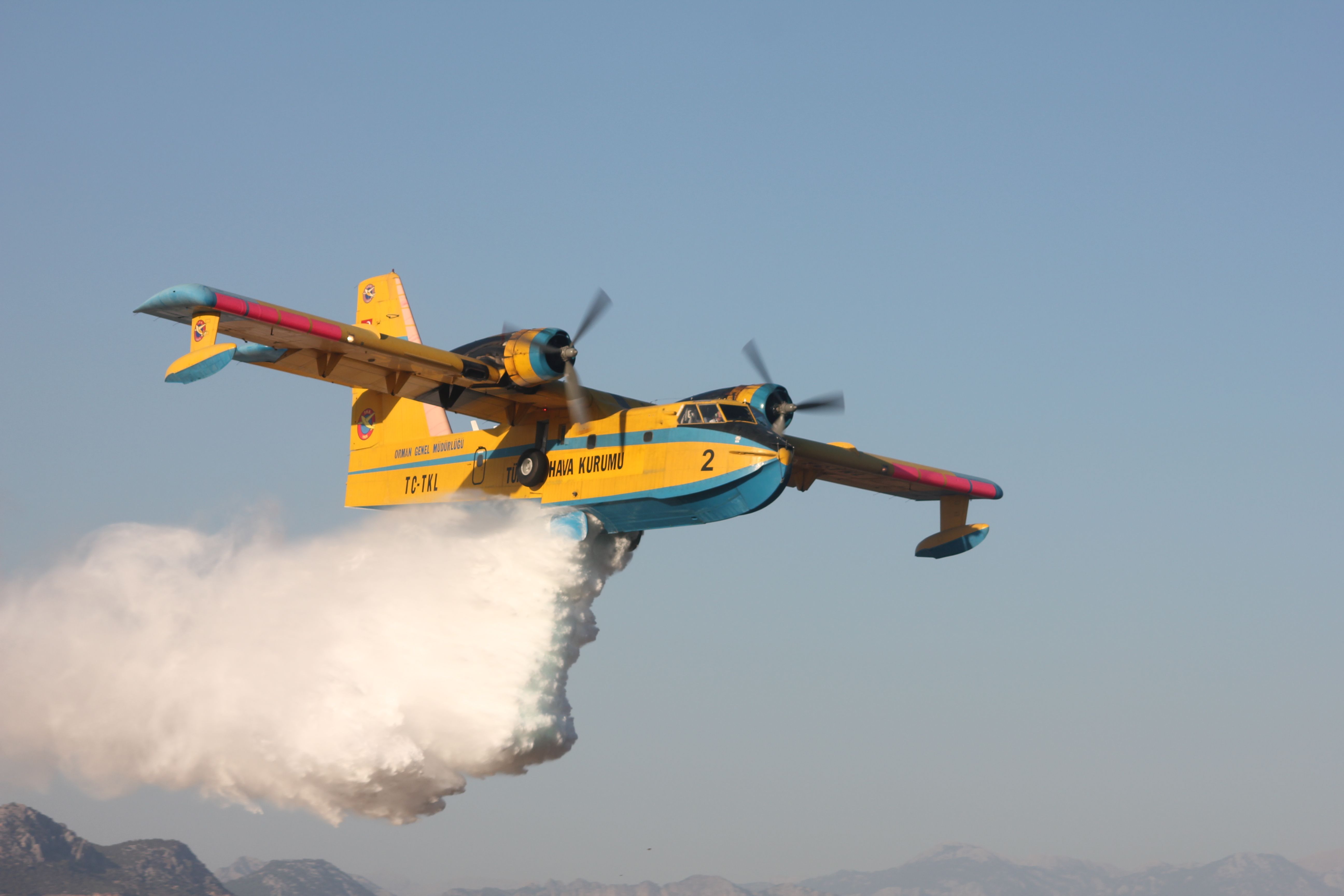 Yellow and blue water bomber plane releases water over mountainous landscape on a clear day.