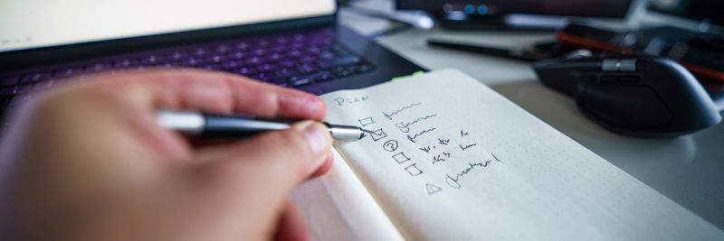 Hand writing in notebook on a desk with laptop, showcasing productivity and organization skills.