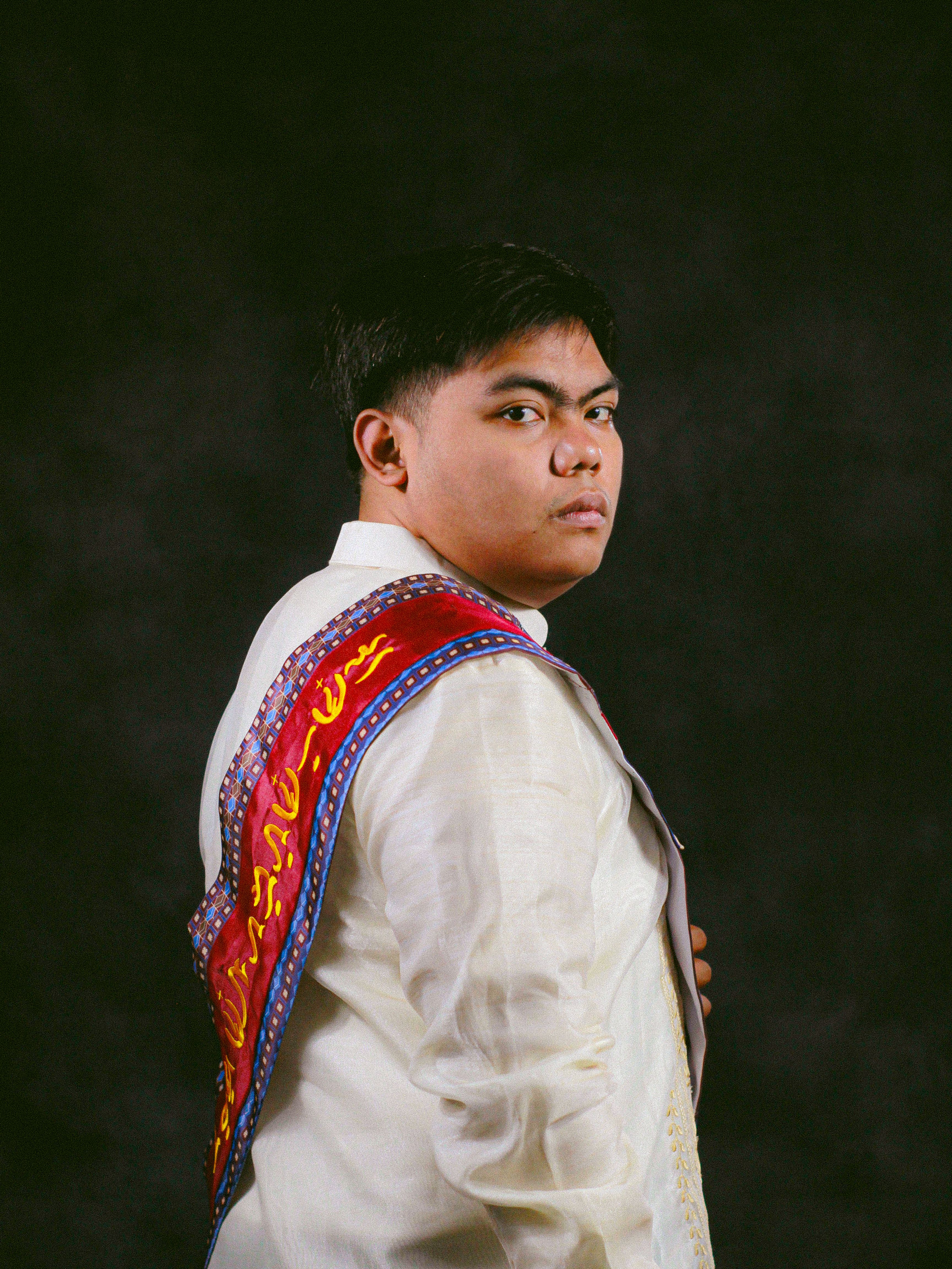 Portrait of a man wearing a traditional barong with a vibrant sash, set against a dark background.