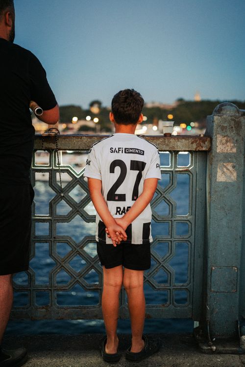 Free A young boy wearing a football jersey stands by the water in Istanbul at dusk. Stock Photo