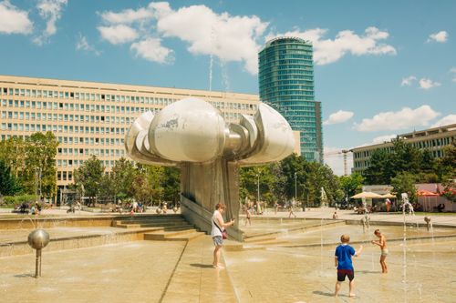 Free Children enjoy a modern urban fountain in a city plaza under a sunny sky, surrounded by buildings. Stock Photo