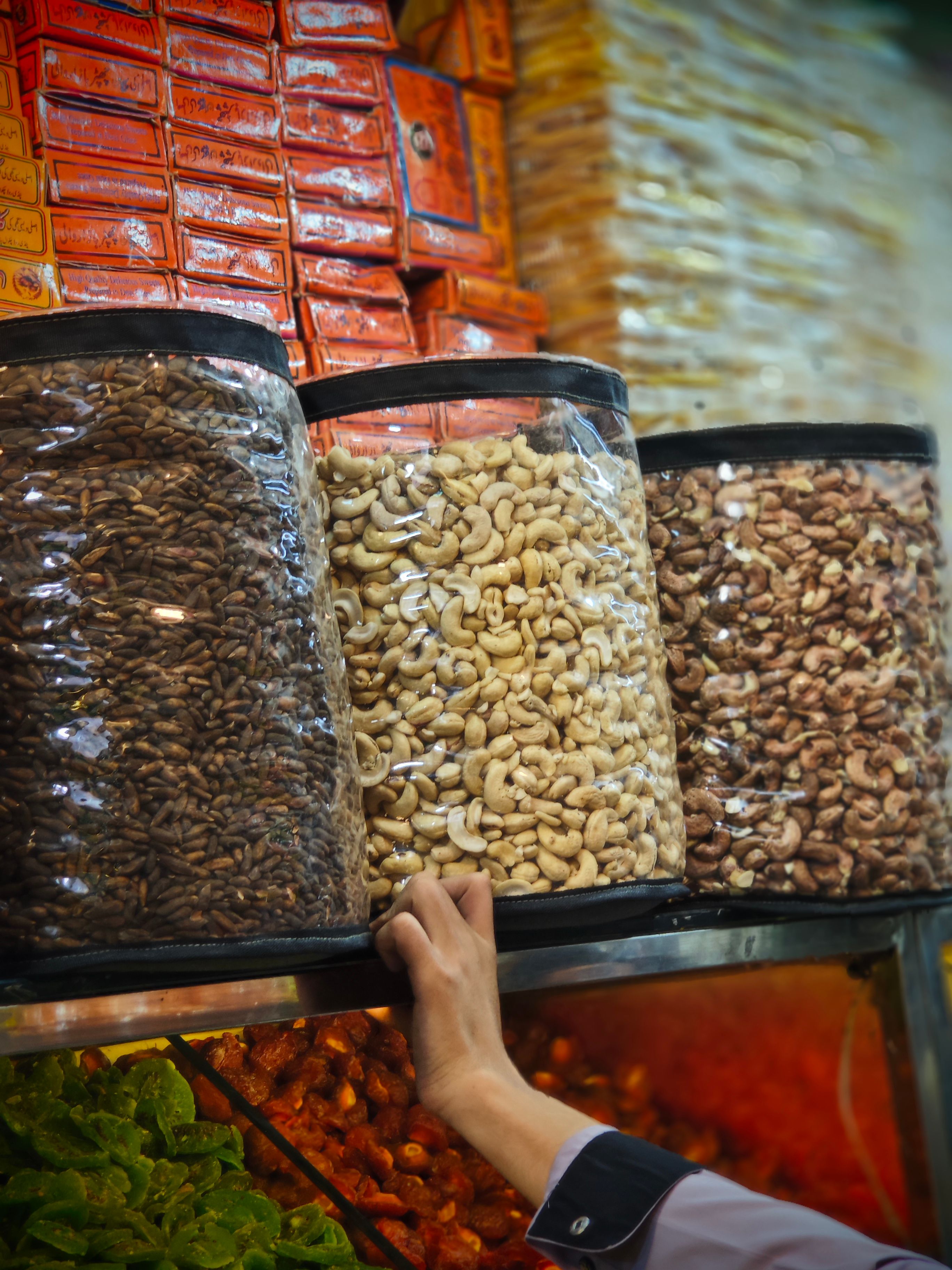 Hand reaching for assorted nuts in jars at a vibrant market stall. - Types of Dry Fruits Available in Pakistan