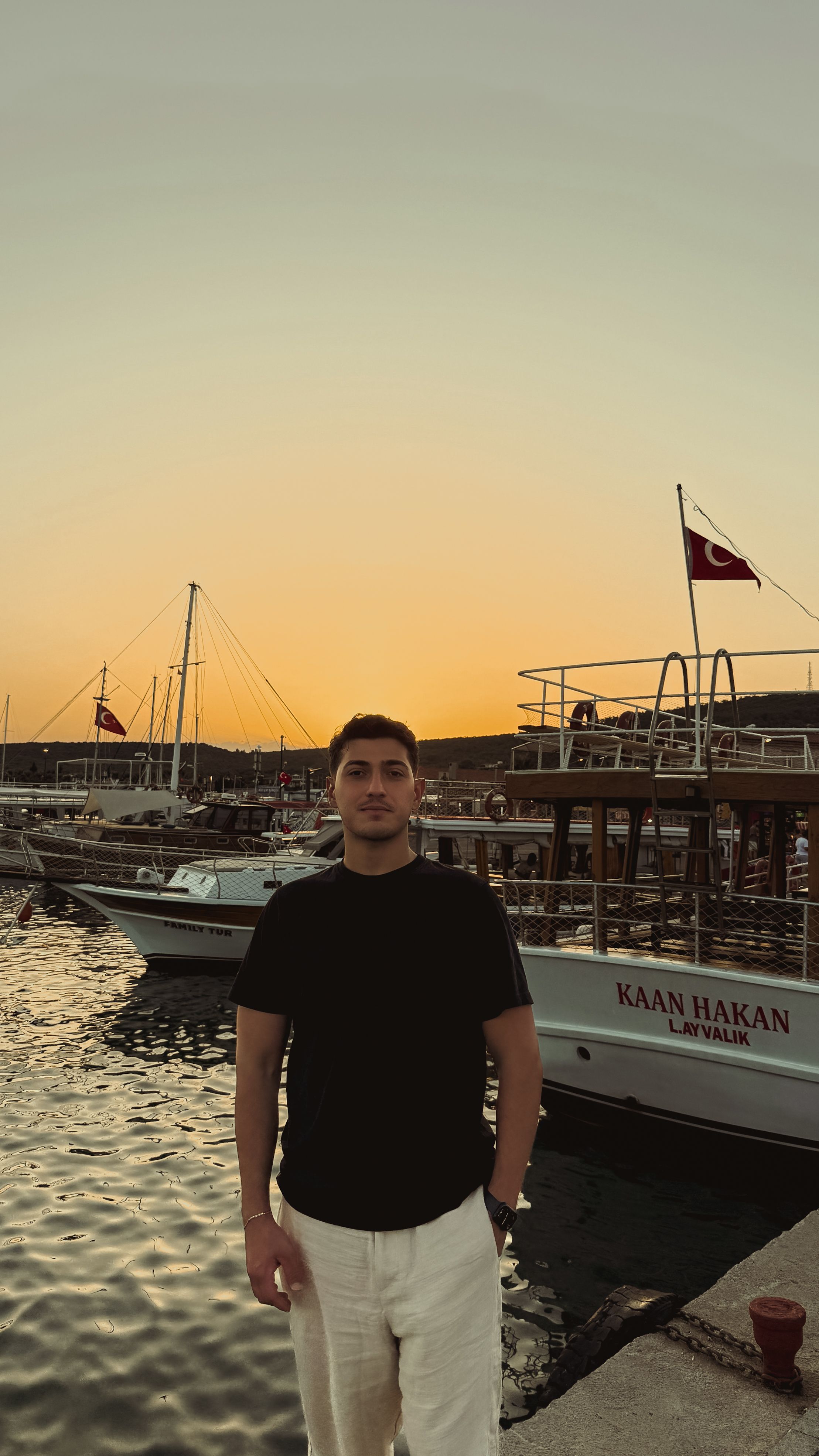 Photograph of a young man standing at Ayvalık Marina with boats during sunset, featuring Turkish flags.
