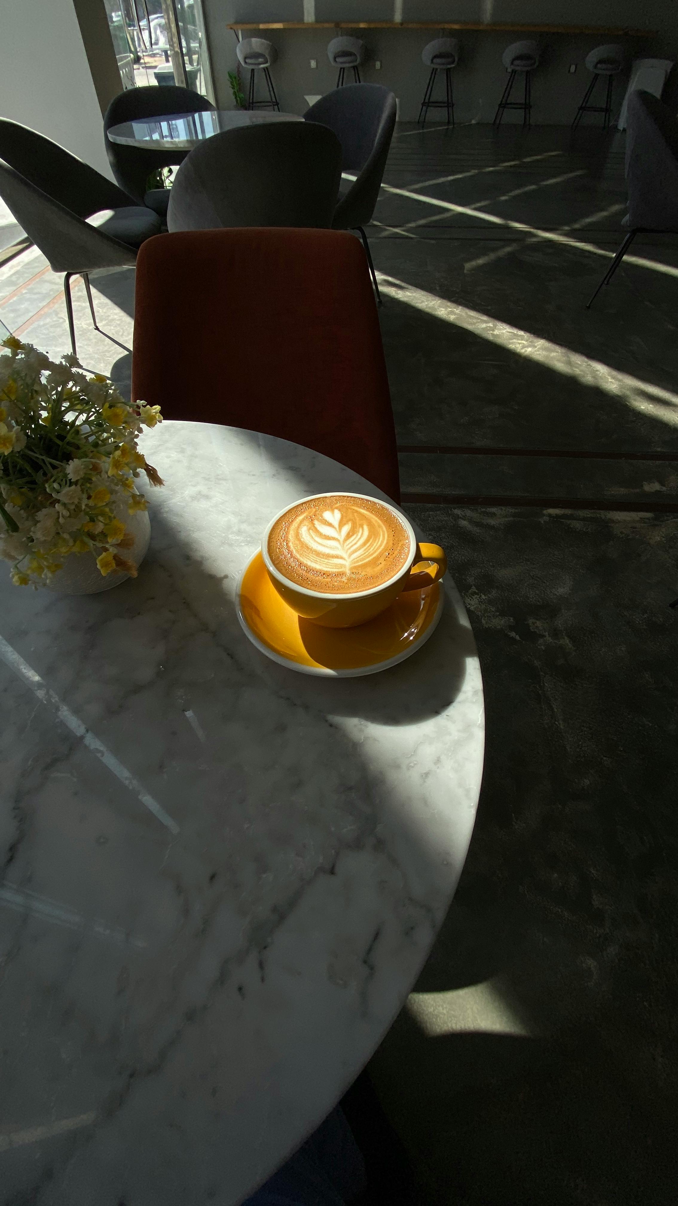 Free A warm cappuccino with latte art sits on a sunlit marble table, surrounded by cozy cafe ambiance. Stock Photo