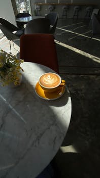 A warm cappuccino with latte art sits on a sunlit marble table, surrounded by cozy cafe ambiance.