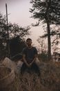 Young sporty ethnic male in casual wear sitting on reinforced cement round shaped product near overgrown grass and trees under serene sky in evening and looking at camera