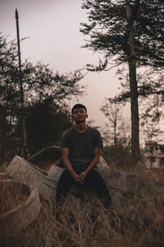 Young sporty ethnic male in casual wear sitting on reinforced cement round shaped product near overgrown grass and trees under serene sky in evening and looking at camera