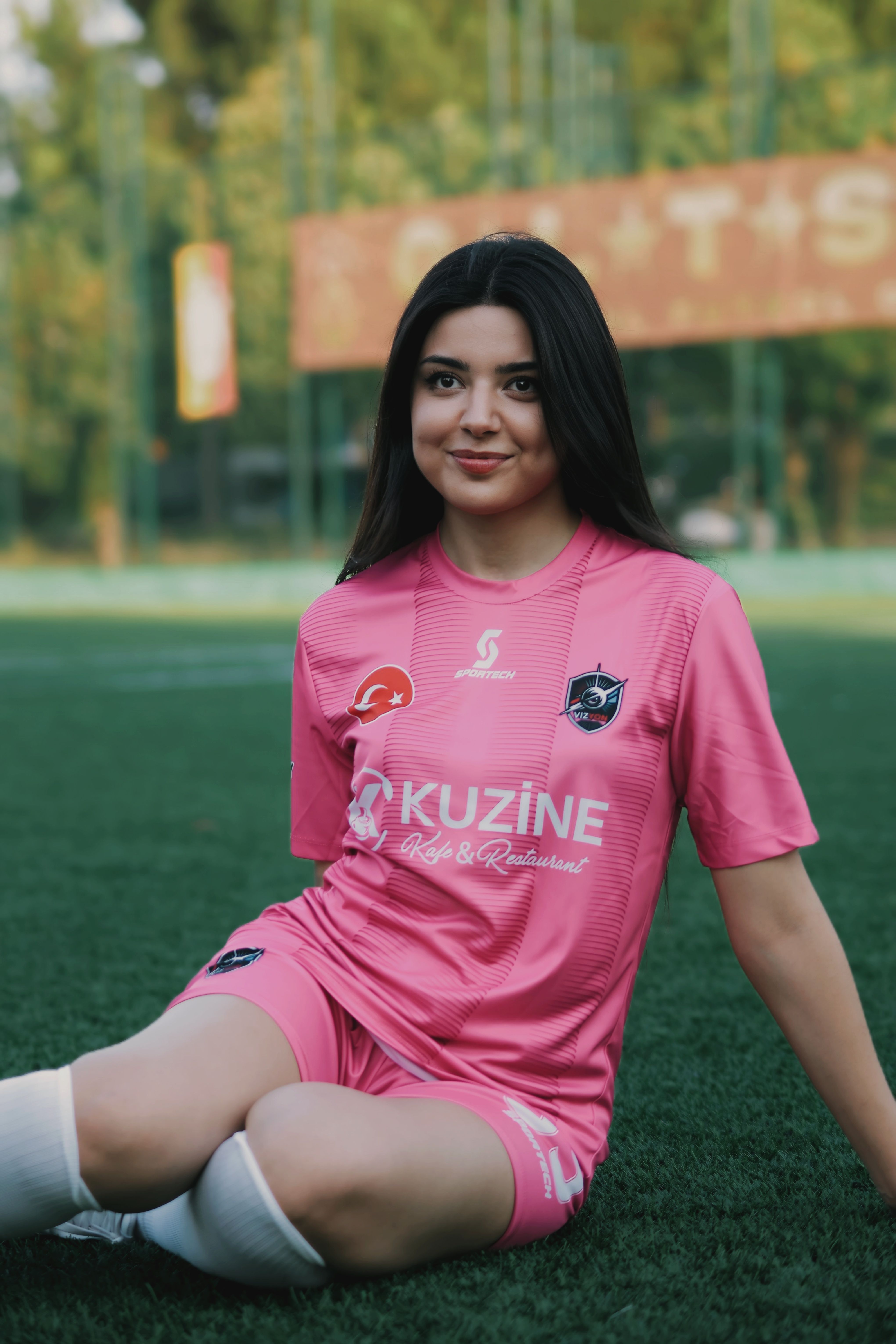 Young woman athlete sitting on a soccer field in a pink uniform posing happily.