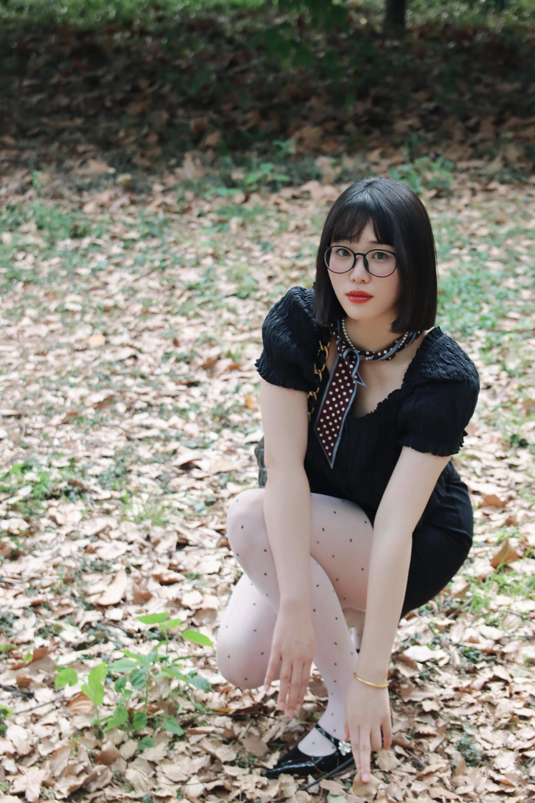 Portrait of a stylish woman crouching outdoors, surrounded by fallen leaves in a serene park setting.