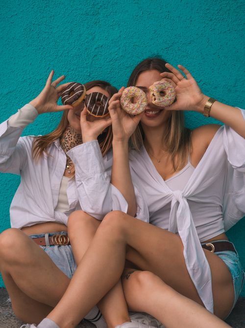 Free Two women laughing with doughnuts by a blue wall, embodying youthful joy and summer fun. Stock Photo