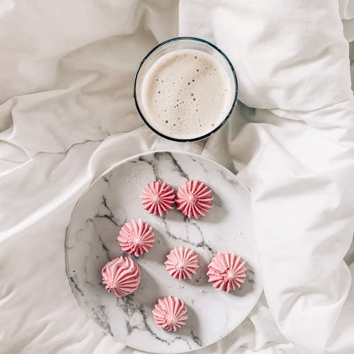 Top view of pink crunchy meringues with ribbed surface on round ceramic plate near glass of hot foamy coffee on creased bed sheet in morning at home