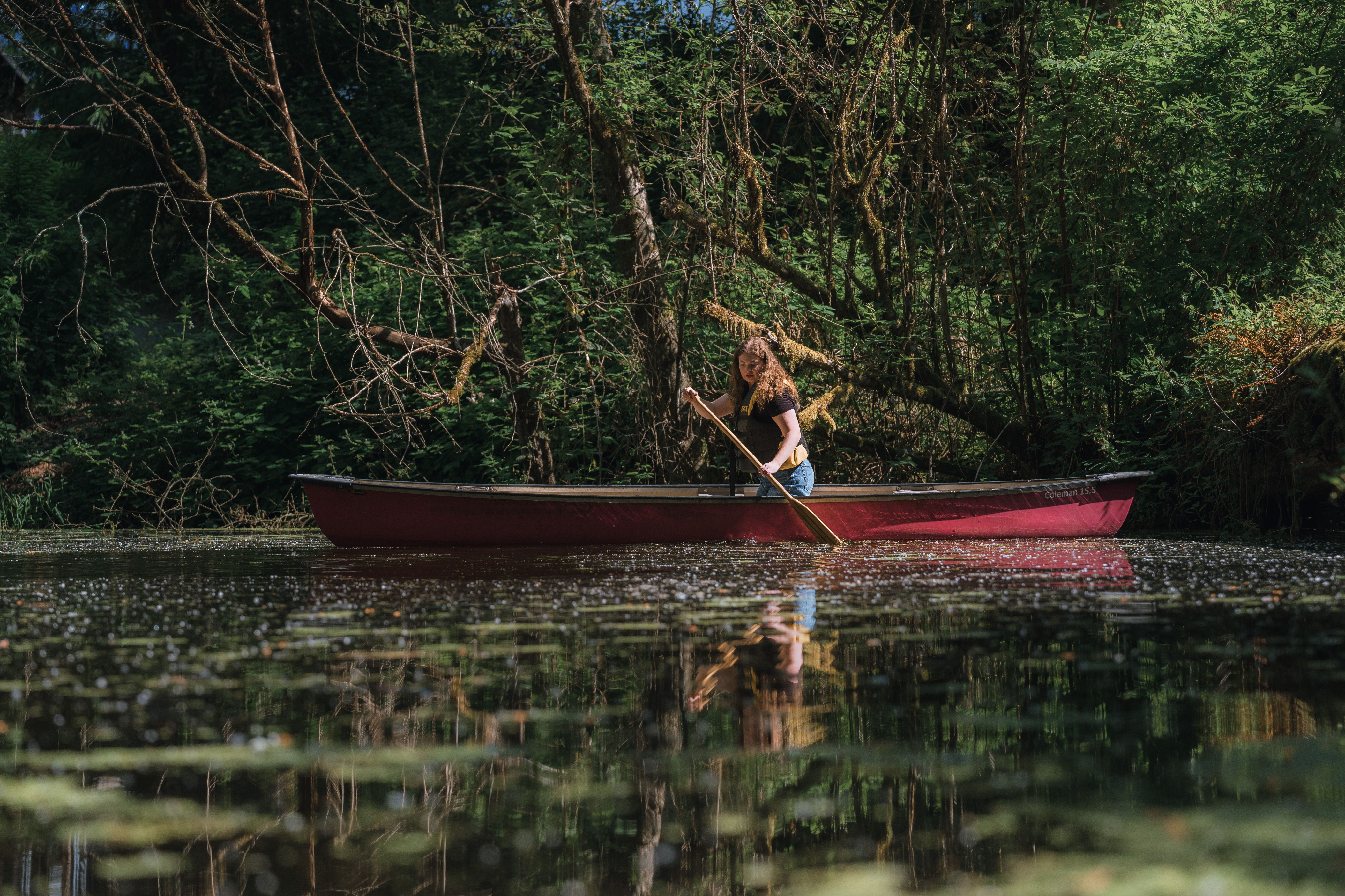 A woman paddles a red canoe across a tranquil, forested lake under dappled daylight.