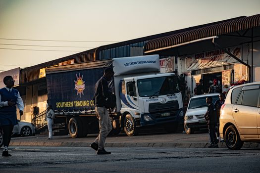 A bustling street scene in Manzini, Eswatini features people and vehicles at twilight.