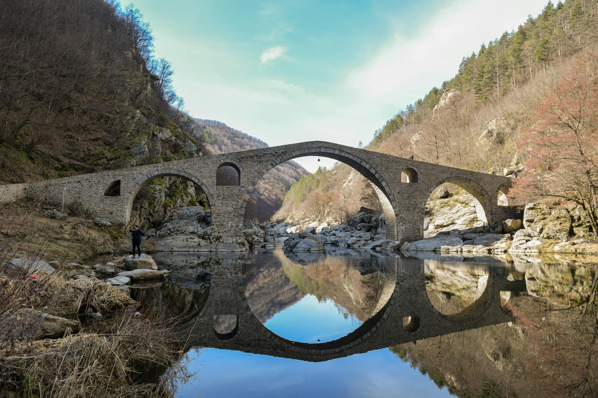 Ancient Devil's Bridge in Bulgaria reflecting in calm river water, framed by lush mountains.