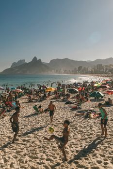Crowded Ipanema Beach in Rio de Janeiro with people playing futvolei under the summer sun.