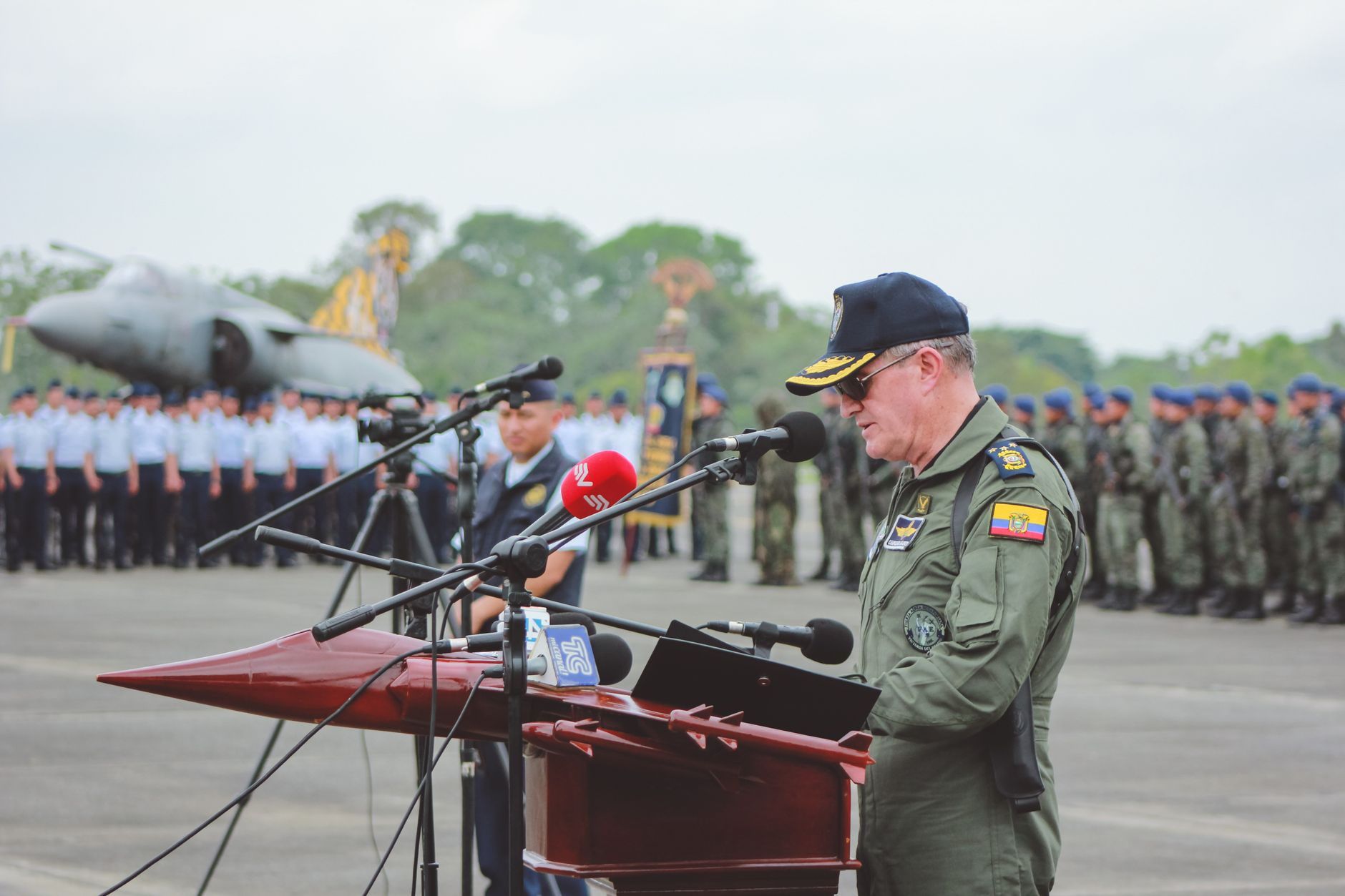 Side view of unrecognizable captain major in sunglasses with shoulder straps speaking into microphone while standing on pavement behind army of soldiers and plane monument during military celebration