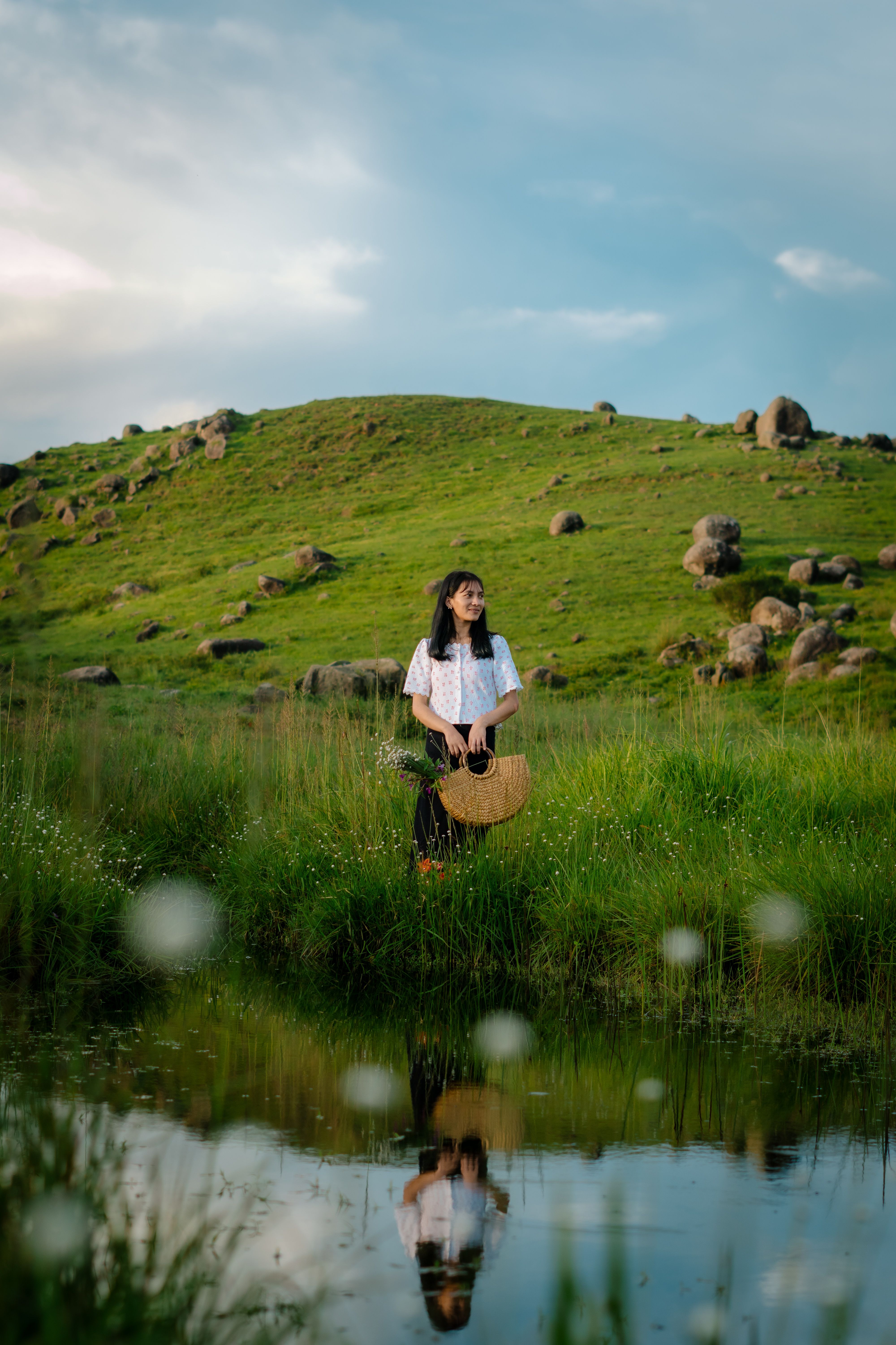 A woman stands by a reflective pond in a lush meadow, ML, India, enjoying a peaceful day.