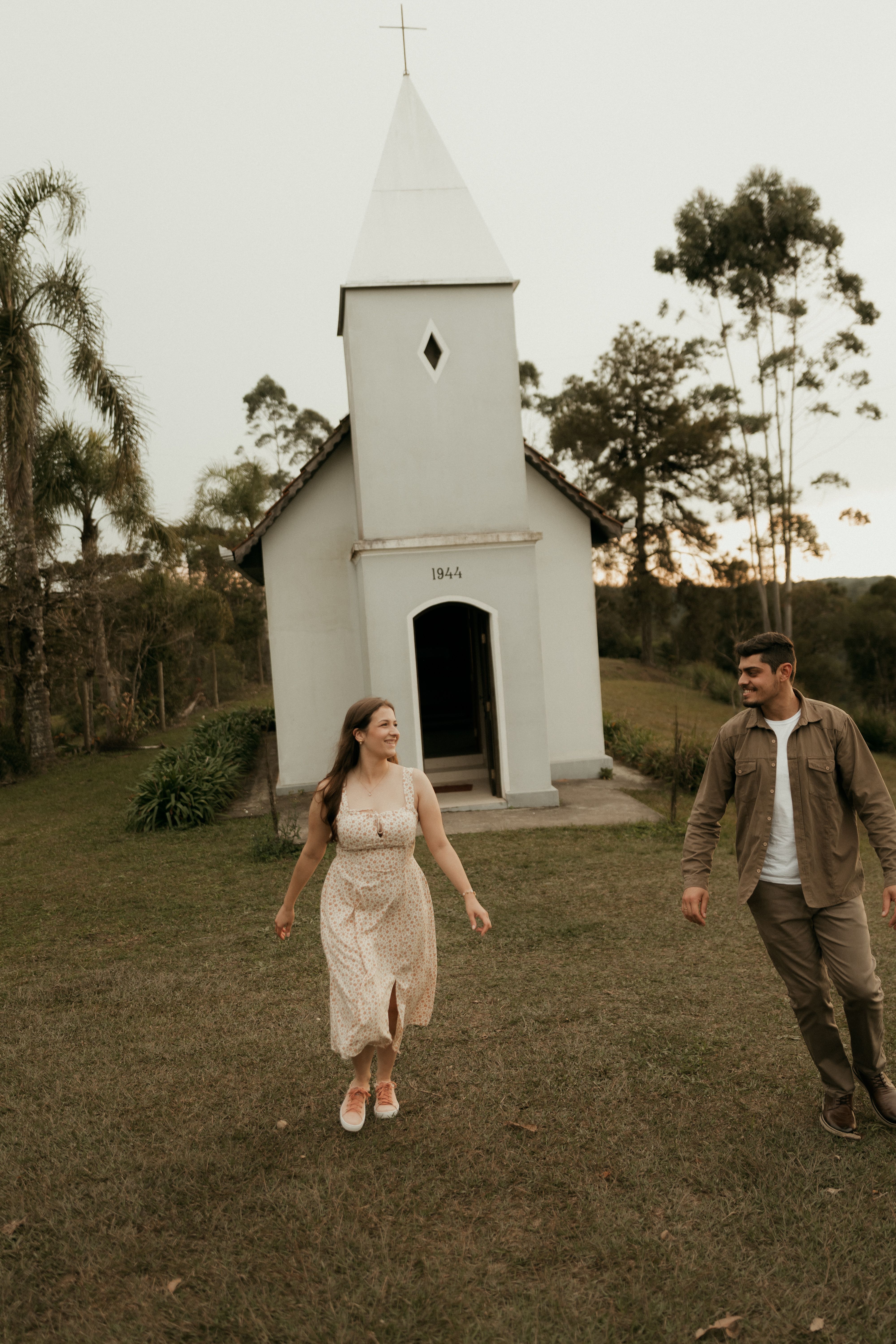 A joyful couple walks hand in hand near a charming small chapel.