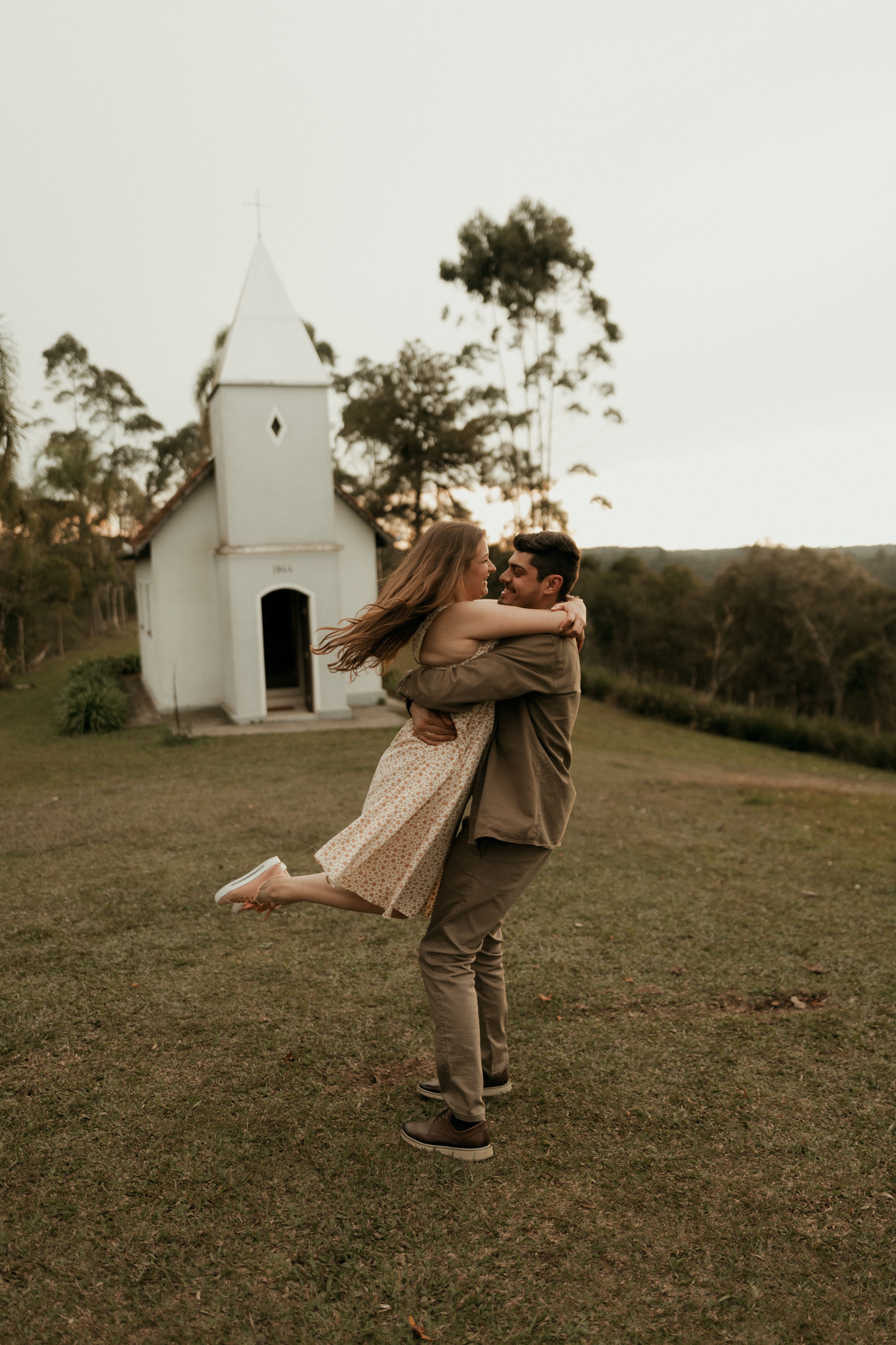 A couple in a loving embrace near a quaint countryside chapel during daylight.