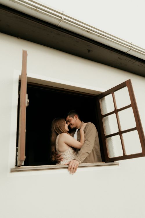 Free A loving couple sharing a tender moment at an open window with a serene background. Stock Photo