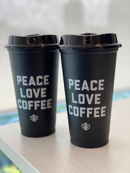 Similar black takeaway plastic glasses with PEACE LOVE COFFEE inscriptions and shiny lids on wooden table for breakfast in morning