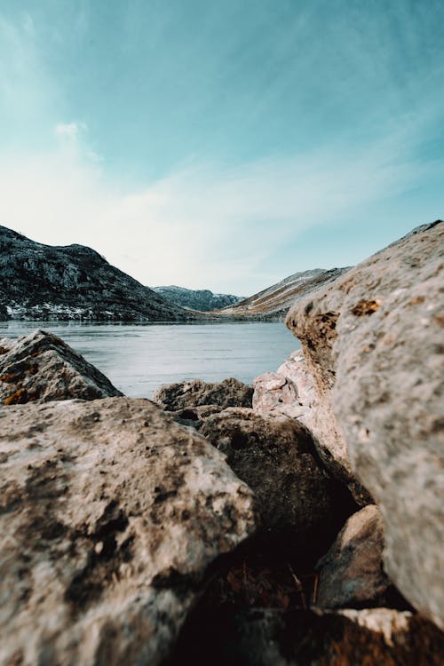 Picturesque view of soundless pond surrounded by high rough mounts and shapeless slopes with uneven surface under bright sky in daylight