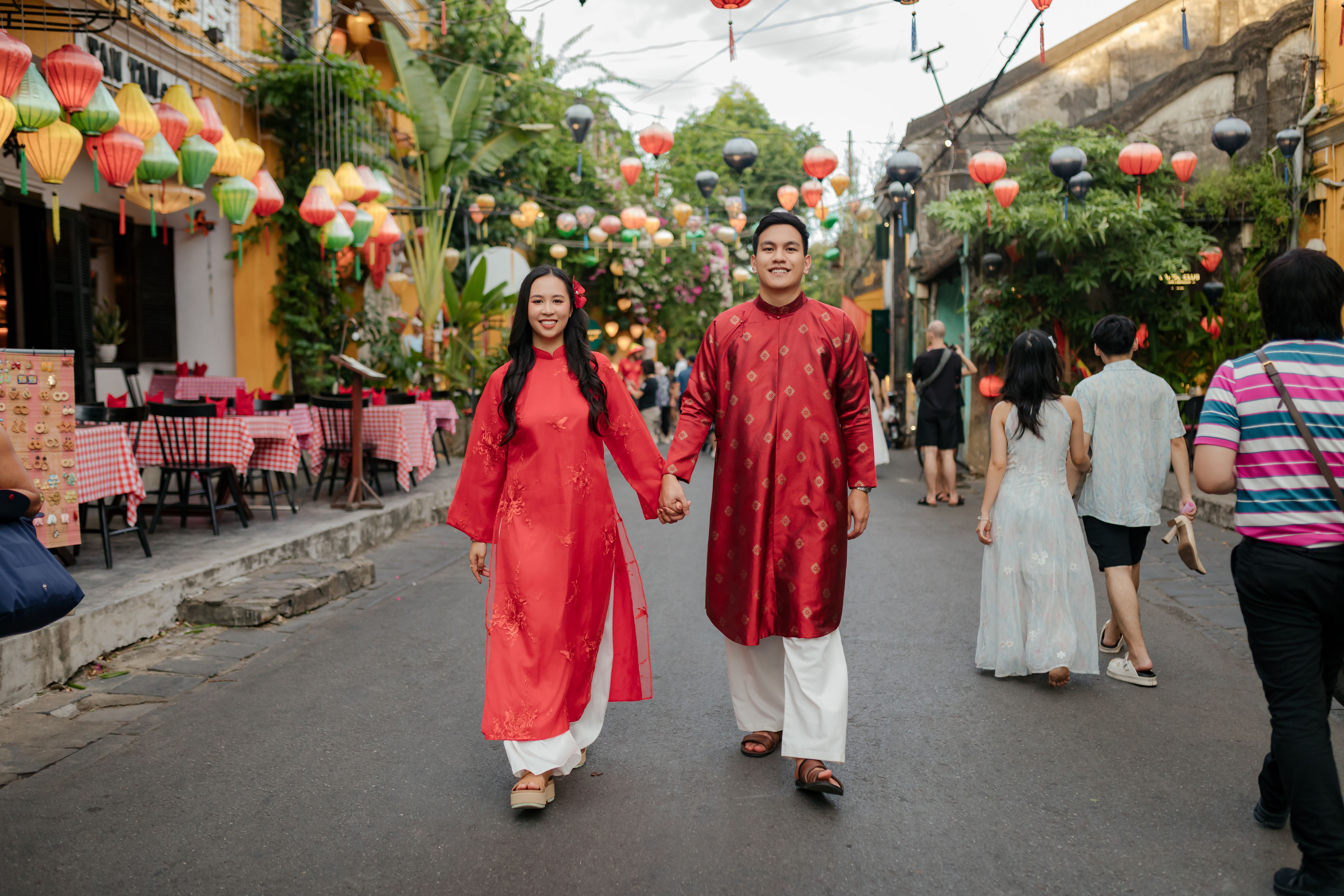 A couple walks hand-in-hand wearing traditional Ao Dai in colorful Đà Nẵng street during daytime.