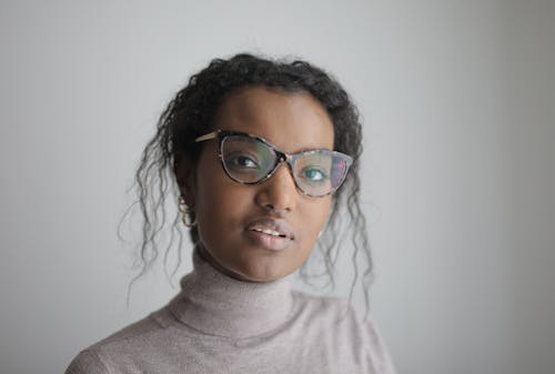 Portrait of young attractive African American female in eyeglasses and casual clothes standing against white wall and looking at camera