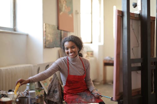 Cheerful smiling African American female in apron sitting on chair in front of easel near table with various tools for painting in creative workshop