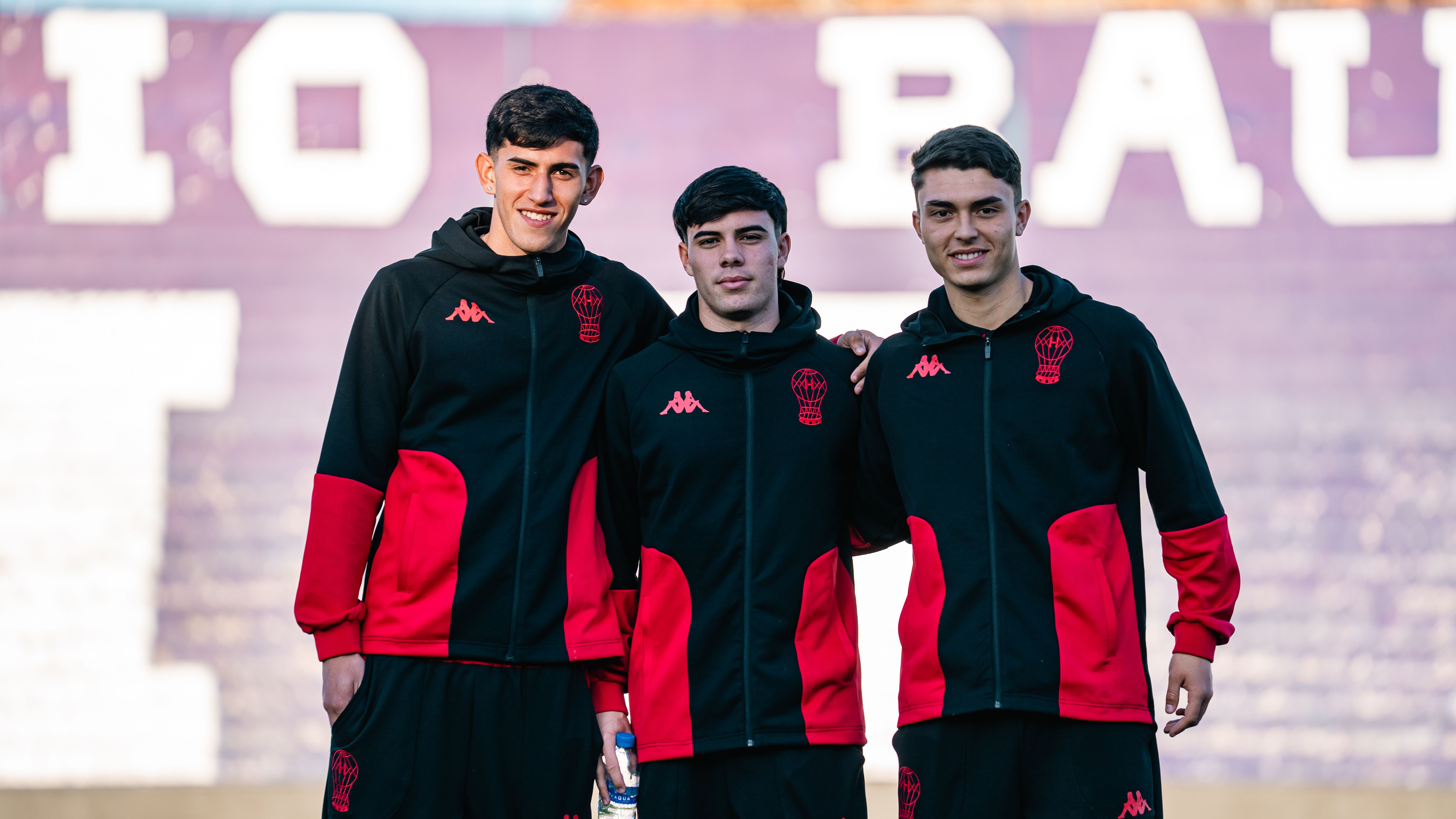 Three young men in sportswear standing outdoors, smiling at the camera.